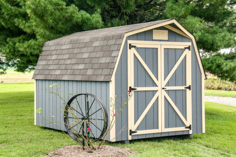 Blue barn-style shed with light beige trim, barn doors, and a shingled roof, located on a grassy yard with trees in the background.