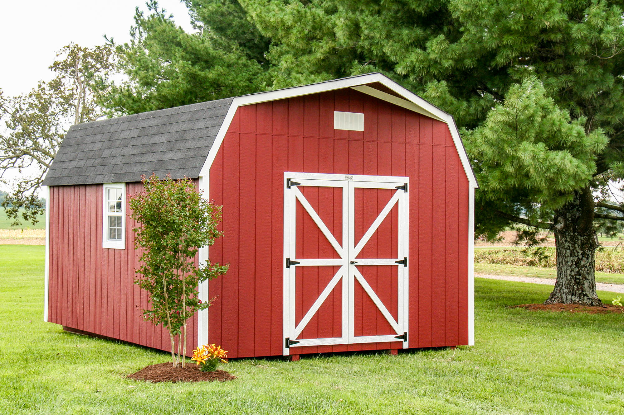 Red barn-style shed with white trim and barn doors, placed on a grassy lawn with small landscaping details