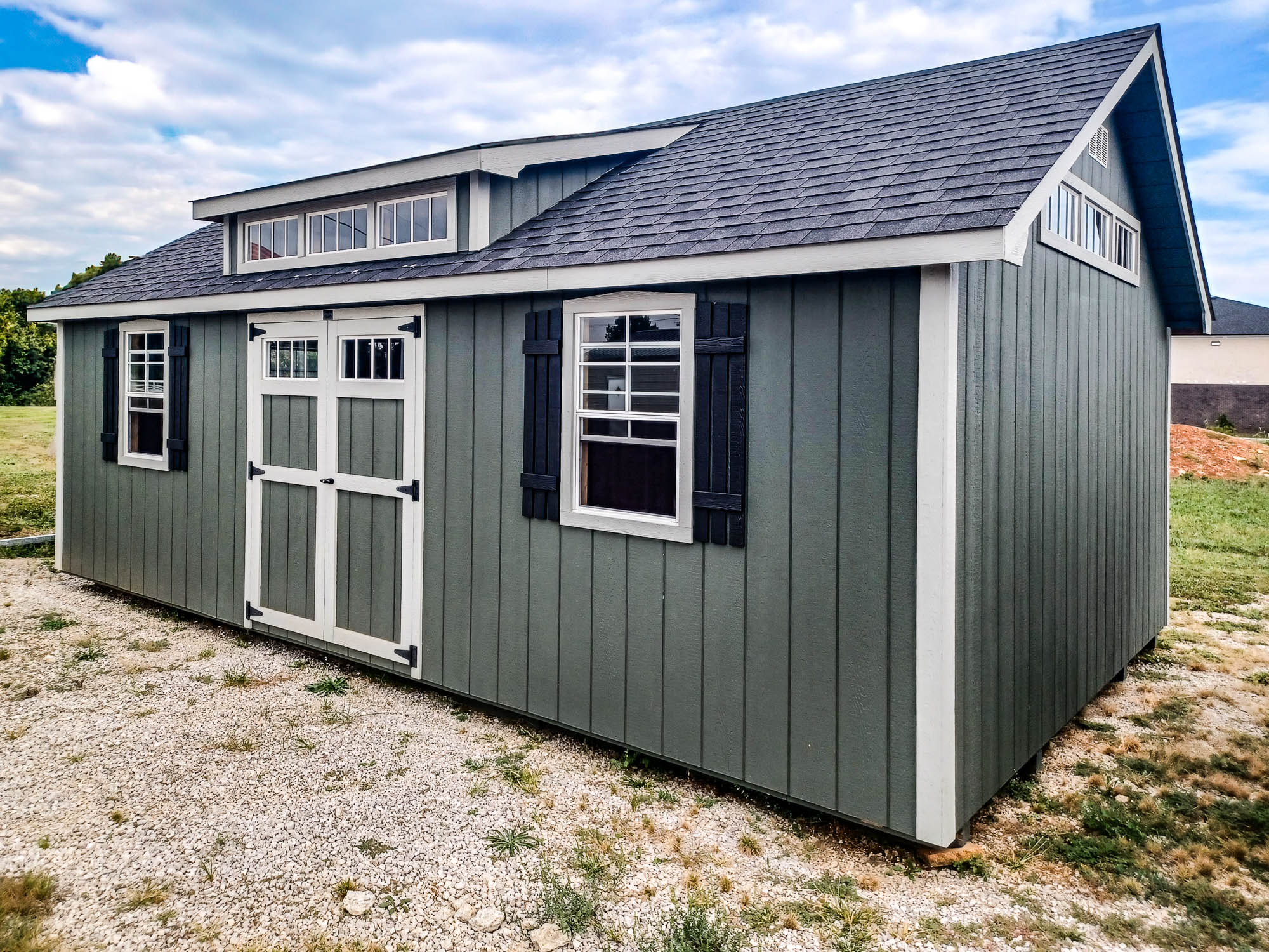 Dark gray shed with white trim, black shutters, and a steeper roof design, featuring a flat central dormer and white-trimmed double doors.