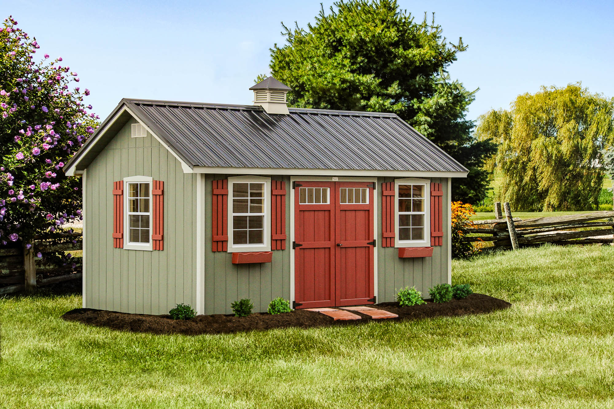 Olive-green shed with red doors, red shutters, and flower boxes, surrounded by landscaping on a sunny lawn.