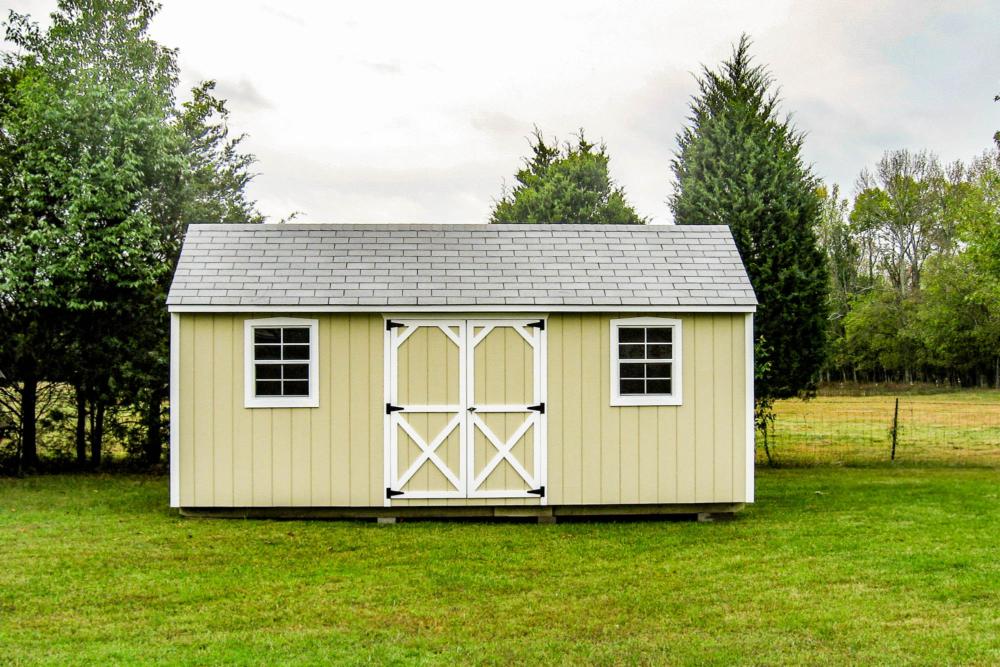 Yellowish beige shed with white trim, barn-style double doors, and two windows, located on a spacious green lawn.