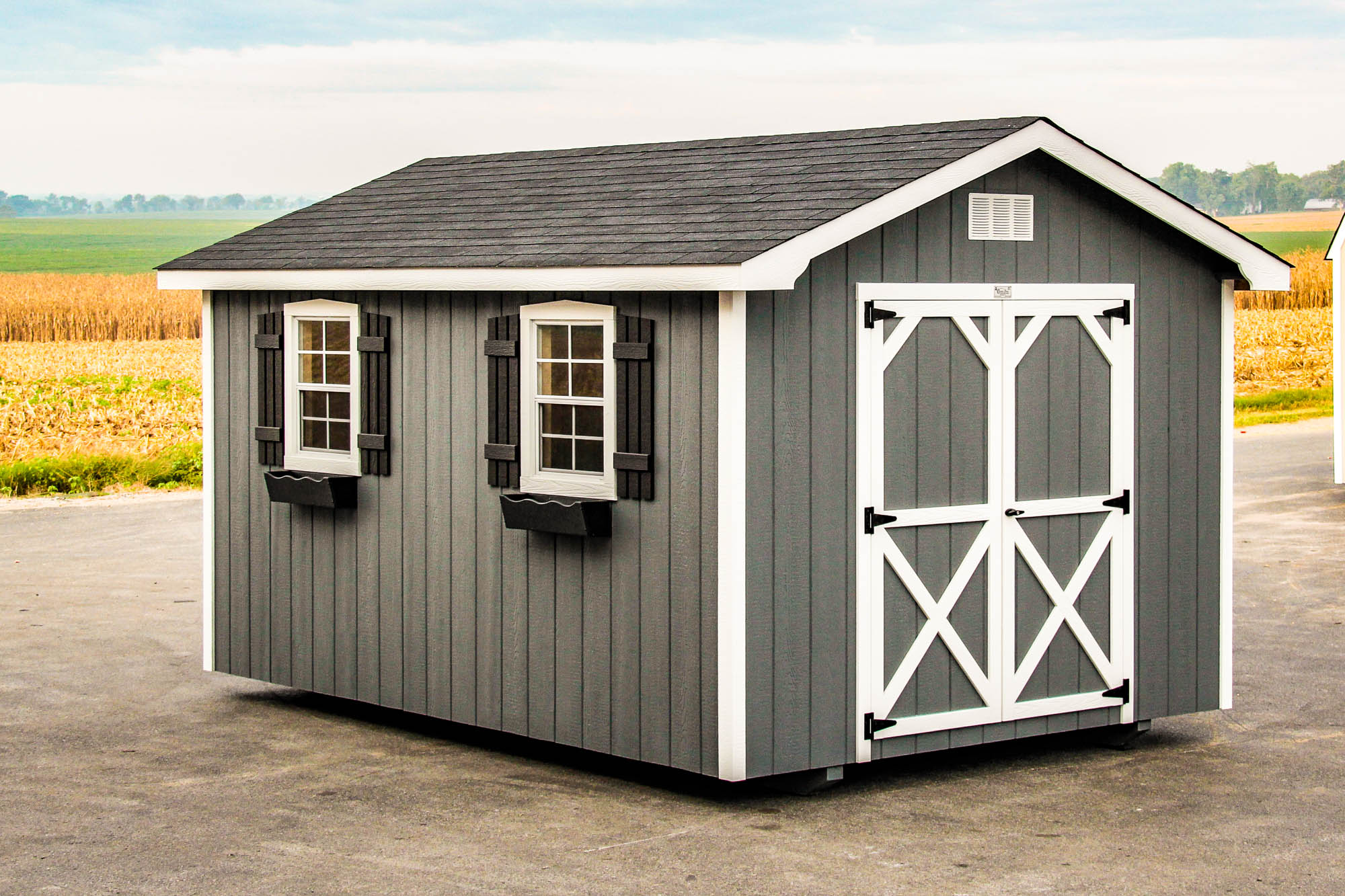 Gray shed with white trim, black shingles, and barn-style doors, set on a paved area with fields in the background.