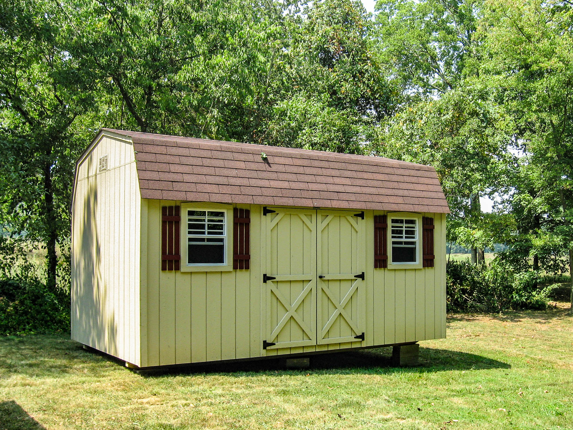 Light tab=n barn-style shed with brown trim and shutters, placed in a shaded backyard with trees.