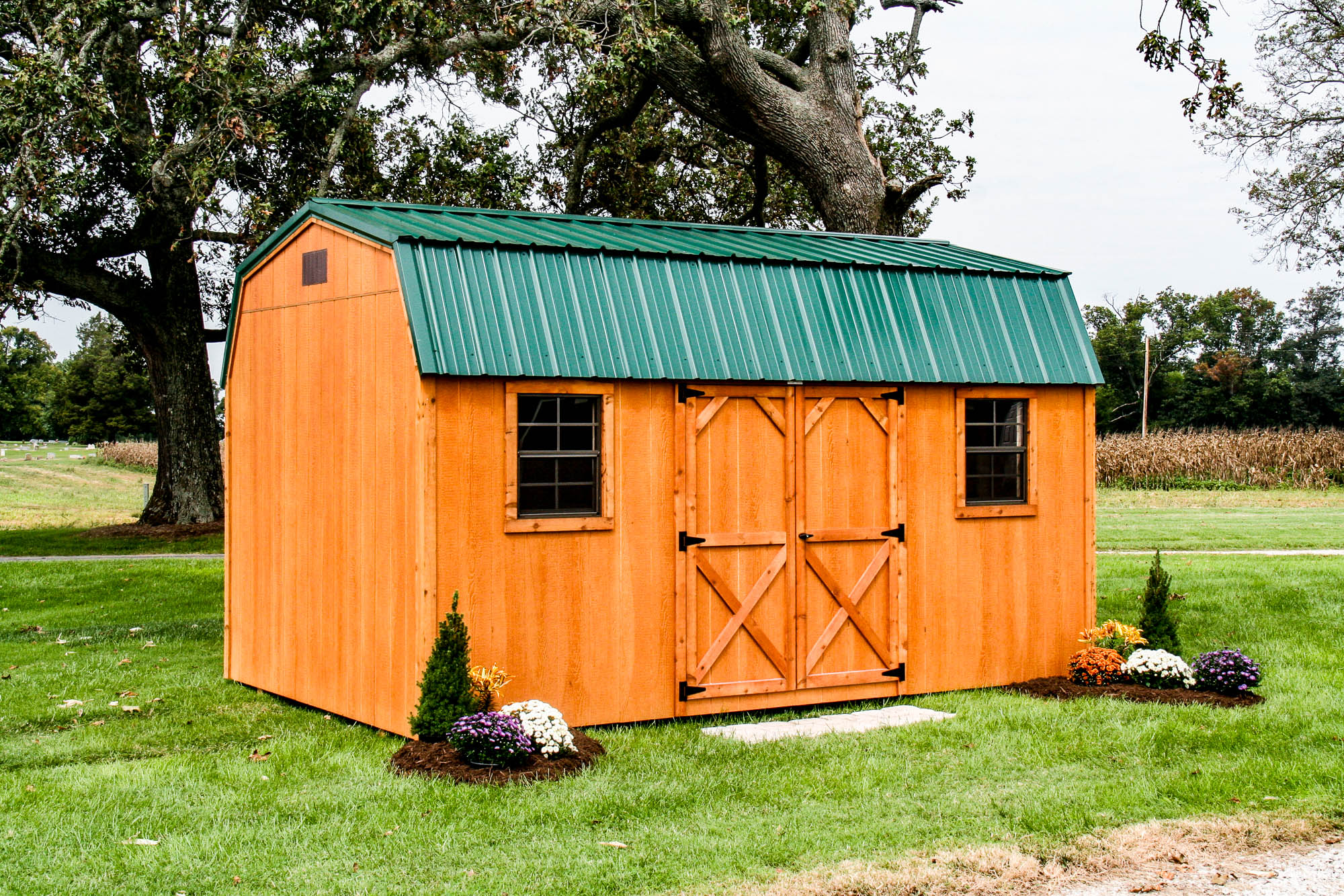 Wooden barn-style shed with a green metal roof, barn doors, and small landscaped flower beds on a grassy lawn.