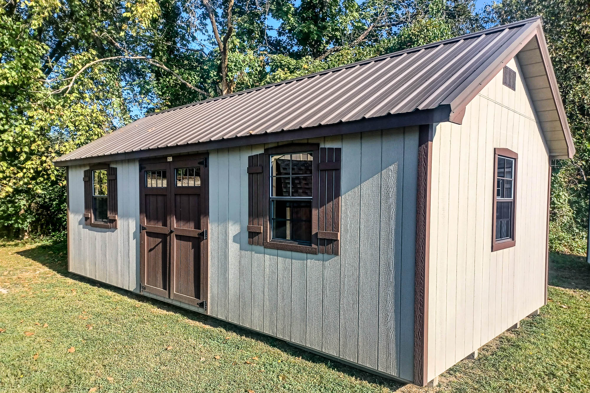 Light beige shed with brown trim, metal roofing, and barn-style doors, located on a grassy yard with trees in the background.