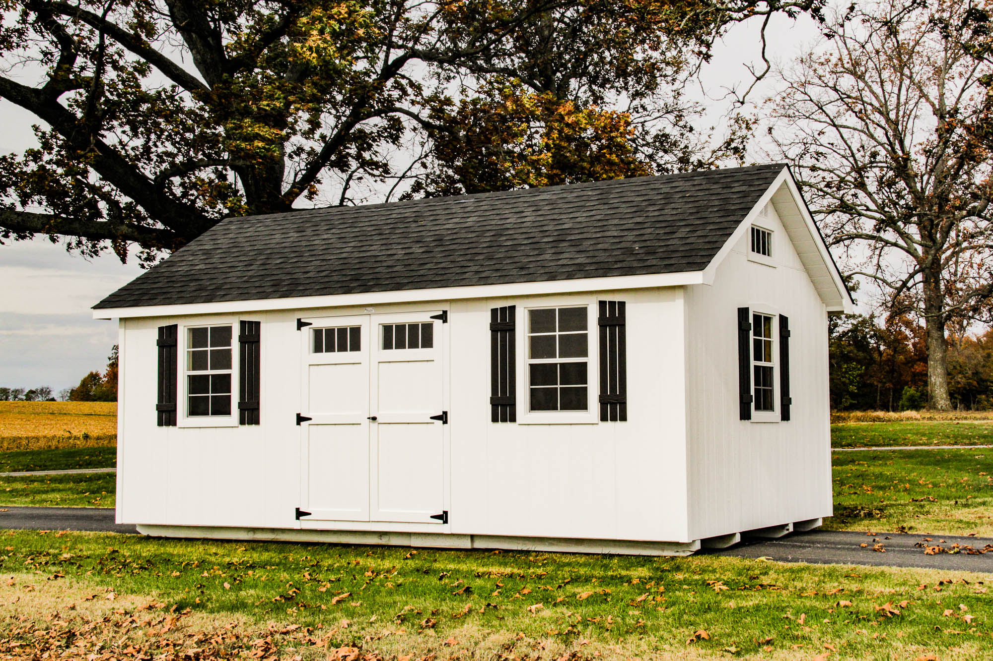 White shed with black shutters and roofing, featuring double doors and windows, surrounded by open fields and trees
