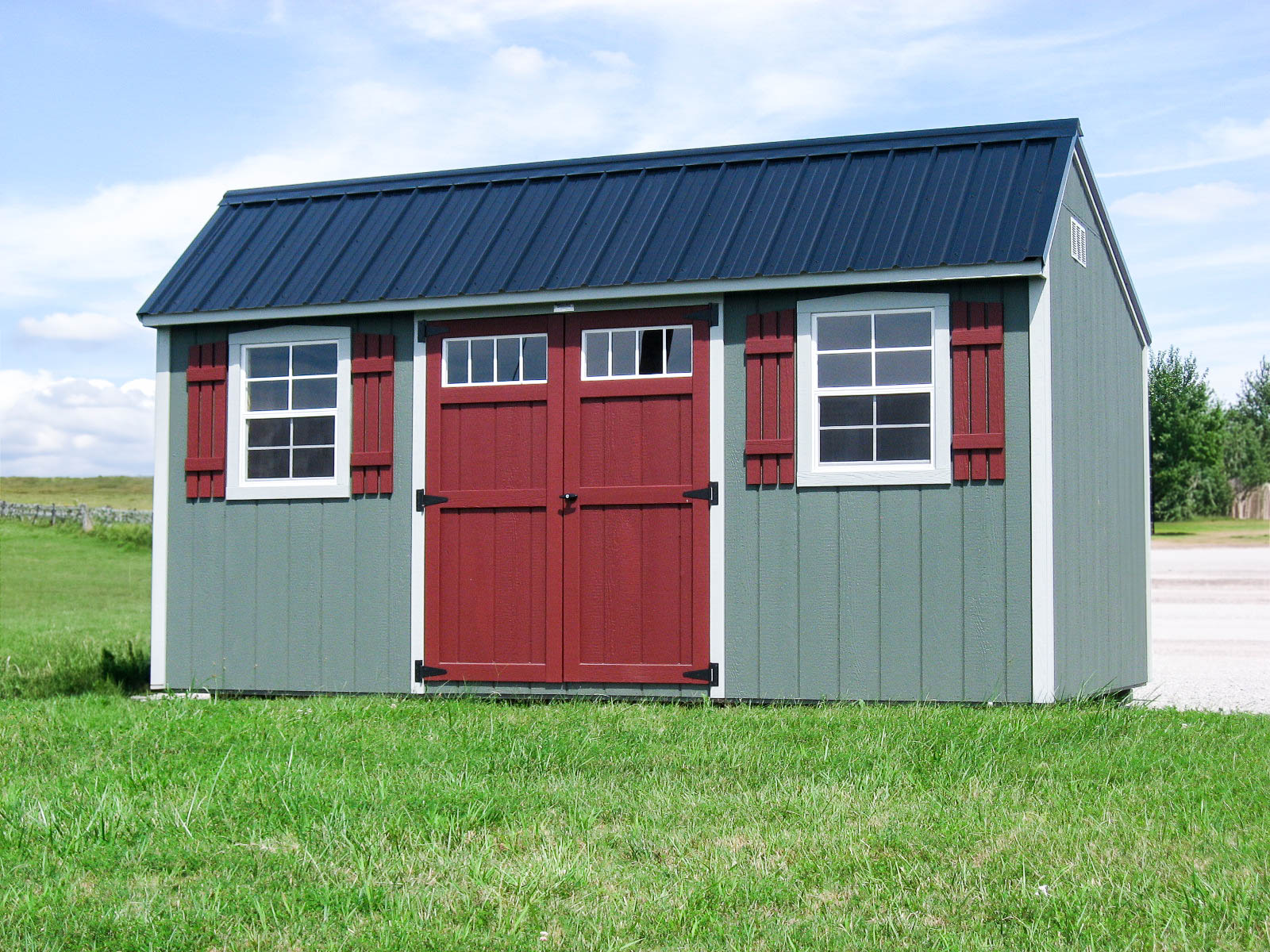 Gray shed with red barn-style doors, red shutters, and black metal roof, placed on a grassy field