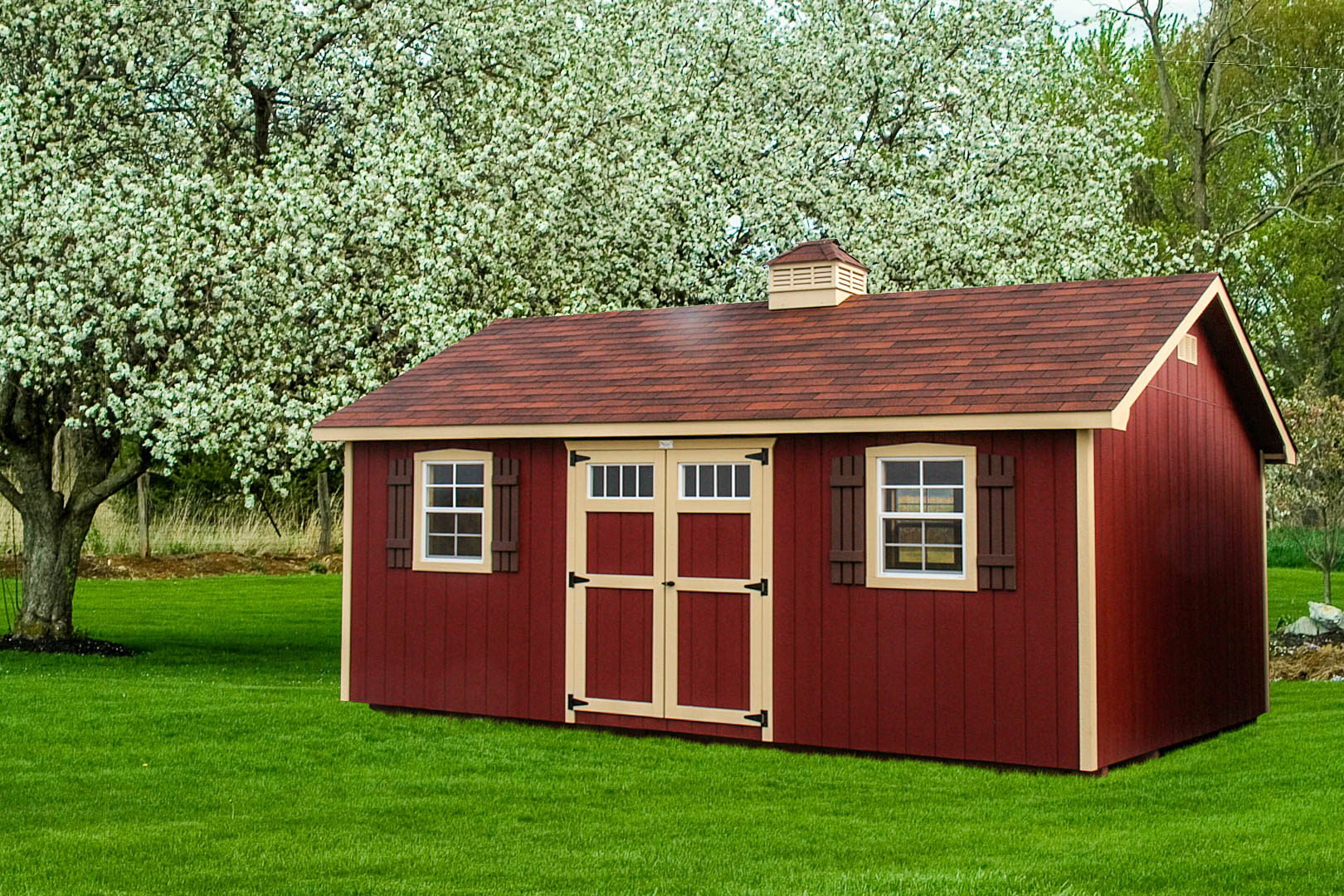Red shed with beige trim, featuring double doors and windows, surrounded by lush greenery."