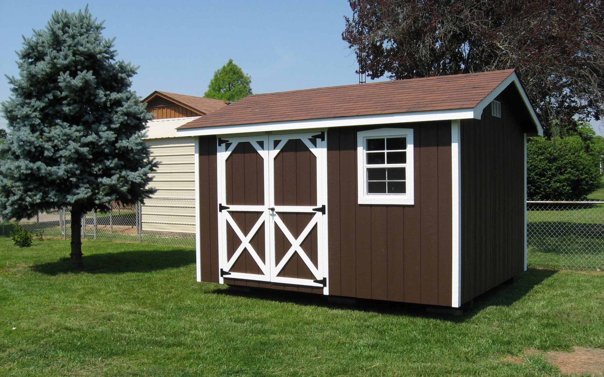 14x32 brown A-roof shed with white trimmed double doors with X detail and a white-framed window to the right - brown roof - shed sits on neat grass near trees and white home