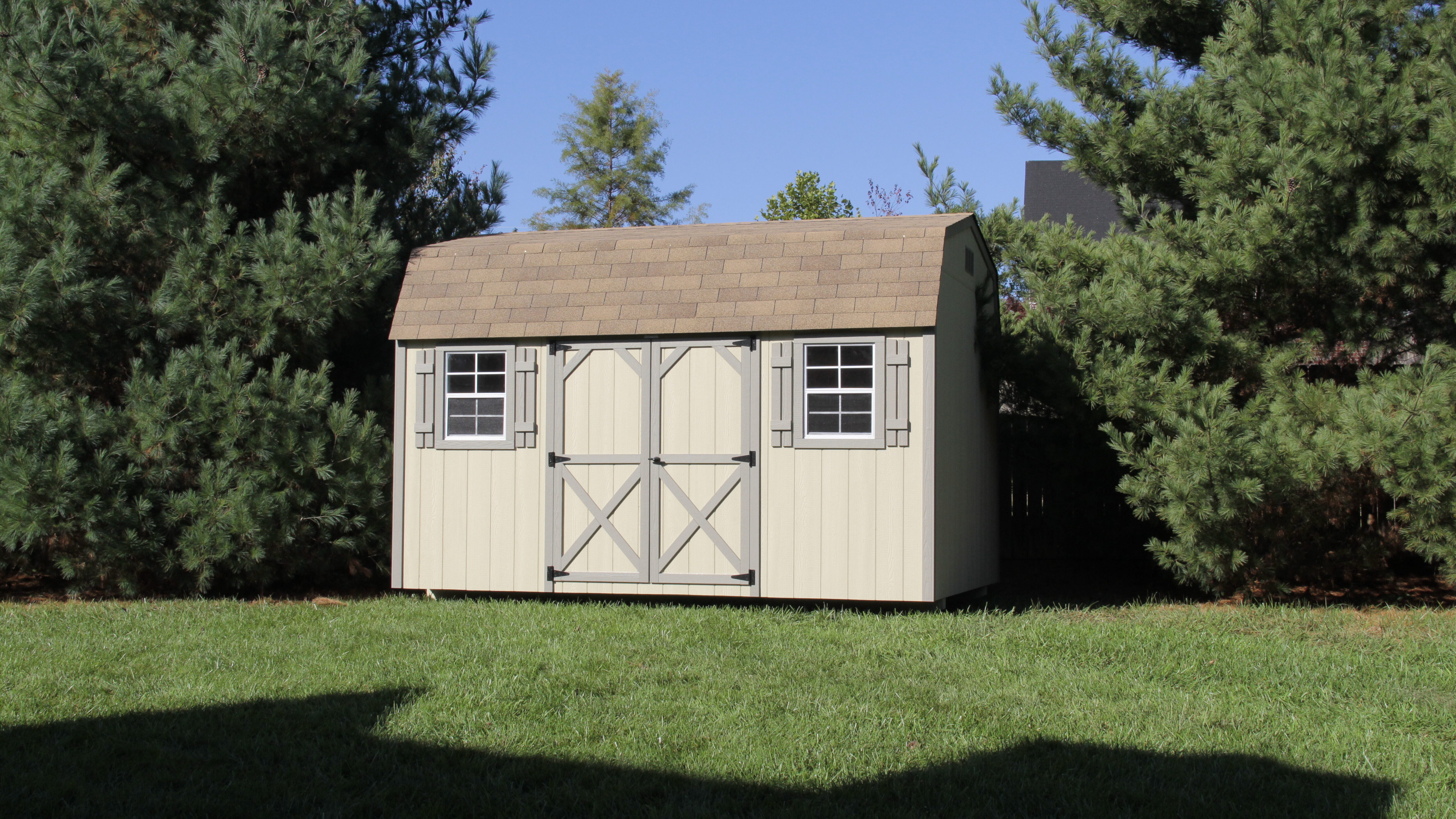 14x32 Deluxe Barn Shed with beige siding and medium brown trimmed double doors between two windows with medium brown shutters beneath brown tiled roof -shed is on green lawn between two trees